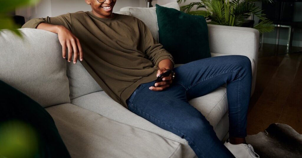 Man setting on the couch looking at his phone smiling and relaxed representing someone who has learned to manage their anxiety effectively through Online Therapy for Anxiety in San Antonio, TX.